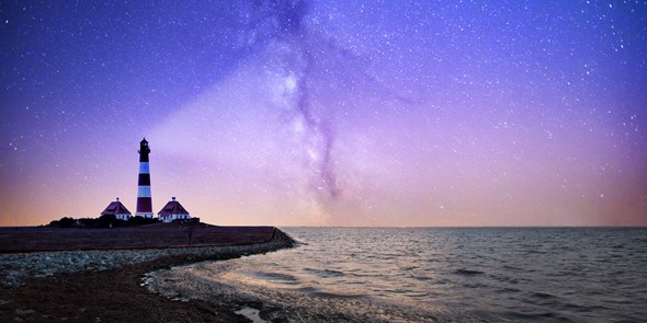 Photograph of a peninsula at nighttime with a lighthouse on the edge of the rocks. The light from the lighthouse projects into the dark sky above which is filled with stars. The sea is calm.