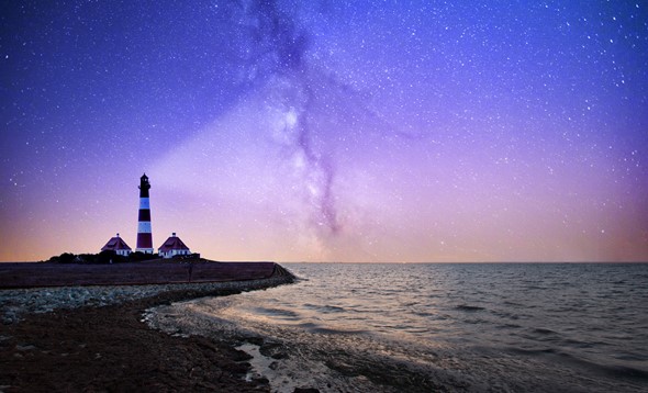 Photograph of a peninsula at nighttime with a lighthouse on the edge of the rocks. The light from the lighthouse projects into the dark sky above which is filled with stars. The sea is calm.