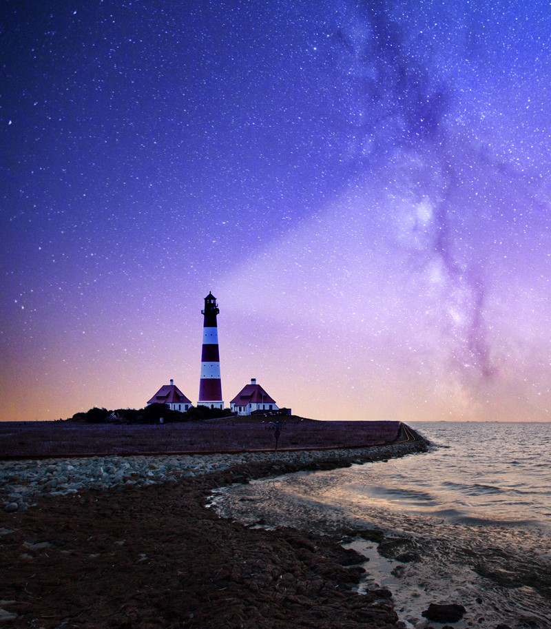 Photograph of a peninsula at nighttime with a lighthouse on the edge of the rocks. The light from the lighthouse projects into the dark sky above which is filled with stars. The sea is calm.