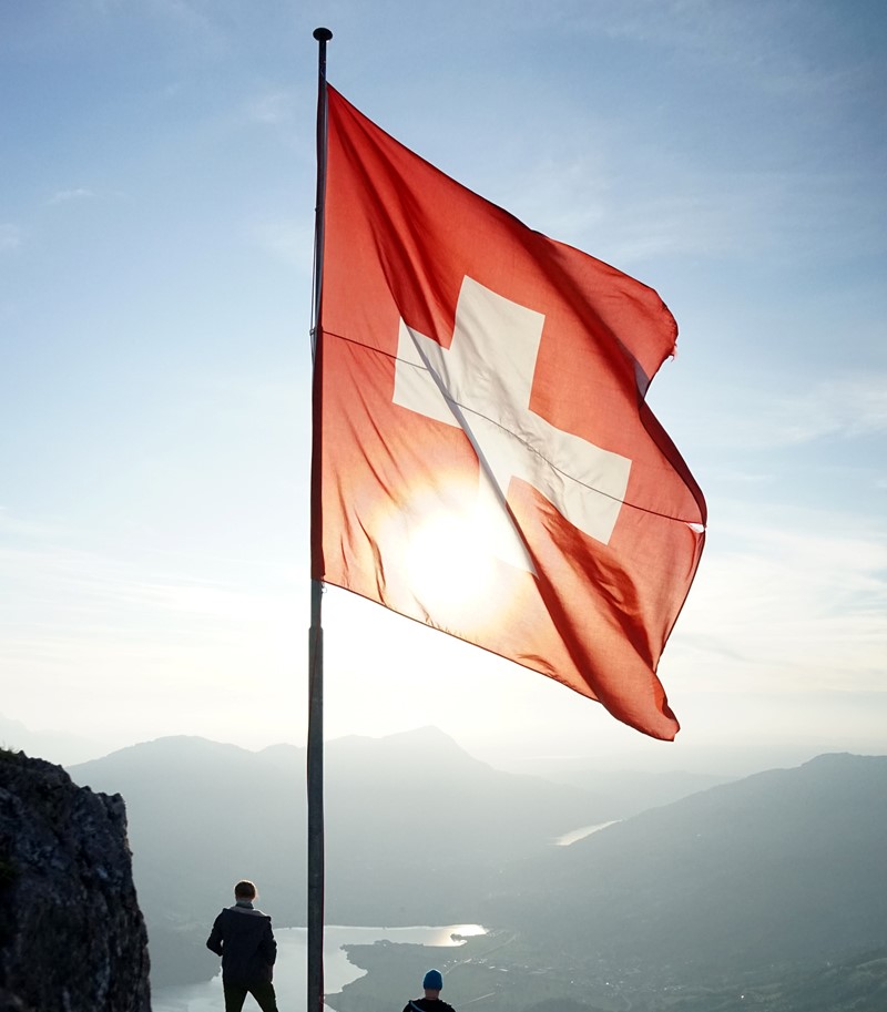 Swiss flag flying from a flag pole on a high vantage point looking out onto a valley and lake below
