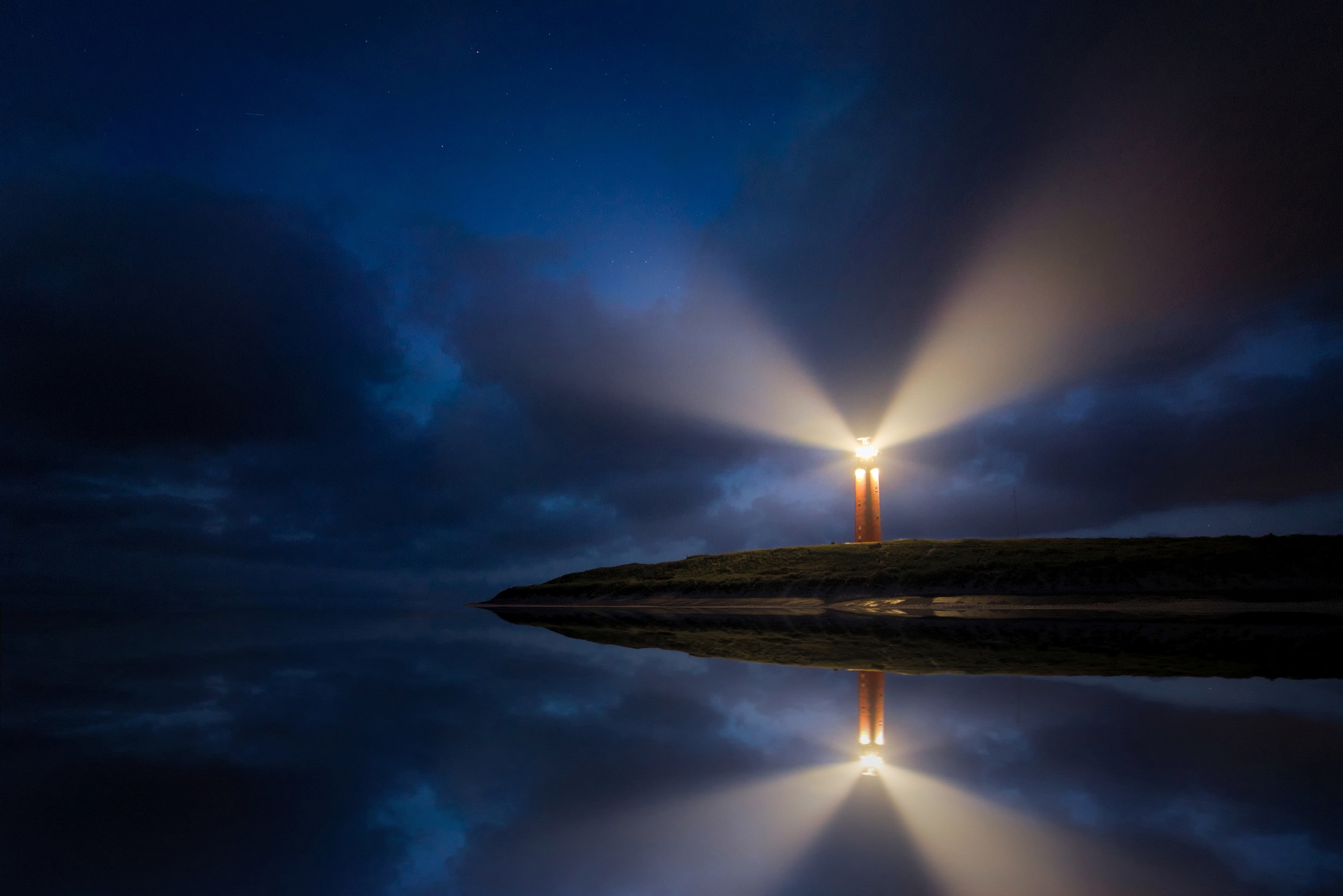Photograph of a peninsula at nighttime with a lighthouse on the edge of the rocks. The light from the lighthouse projects into the dark sky above and is reflected in the calm, still water below. 