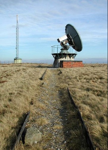 An overgrown path in a grassy field leads to an apparently derelict structure with a navigational dish.
