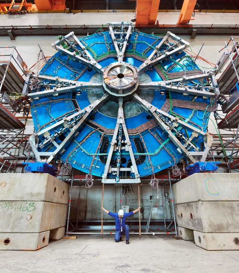 Man in protective gear for building sites kneeling before viewer under a large piece of equipment from CERN