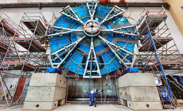 Man in protective gear for building sites kneeling before viewer under a large piece of equipment from CERN
