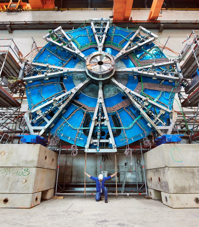 Man in protective gear for building sites kneeling before viewer under a large piece of equipment from CERN