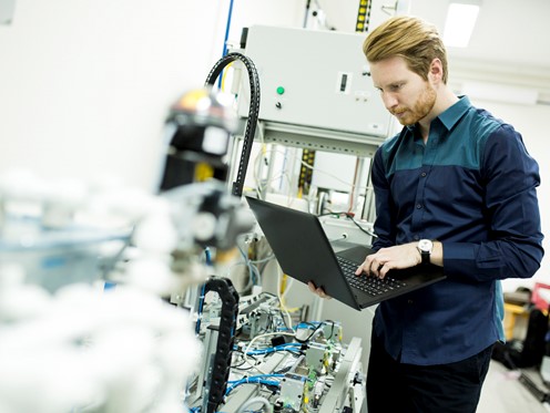 Man with laptop in hand examining complex piece of machinery