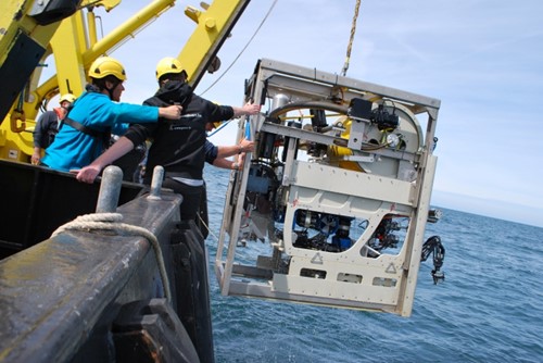 A cube-shaped robot is lifted from the sea on board a ship using a crane