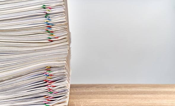 Large stack of files with multicoloured paperclips attached on a wooden desk, stack aligned to the left of the picture