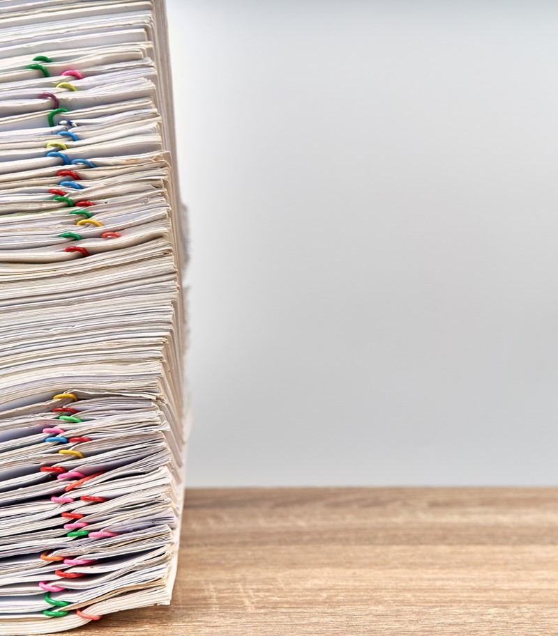 Large stack of files with multicoloured paperclips attached on a wooden desk, stack aligned to the left of the picture