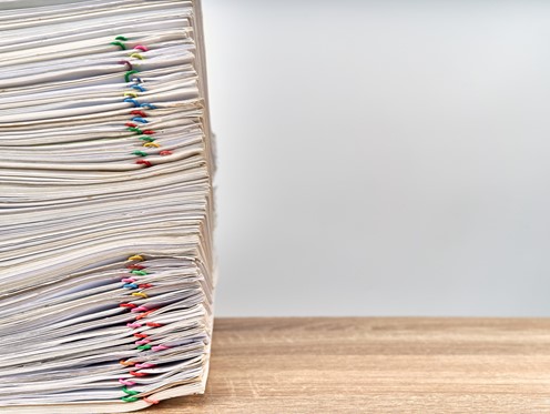 Large stack of files with multicoloured paperclips attached on a wooden desk, stack aligned to the left of the picture